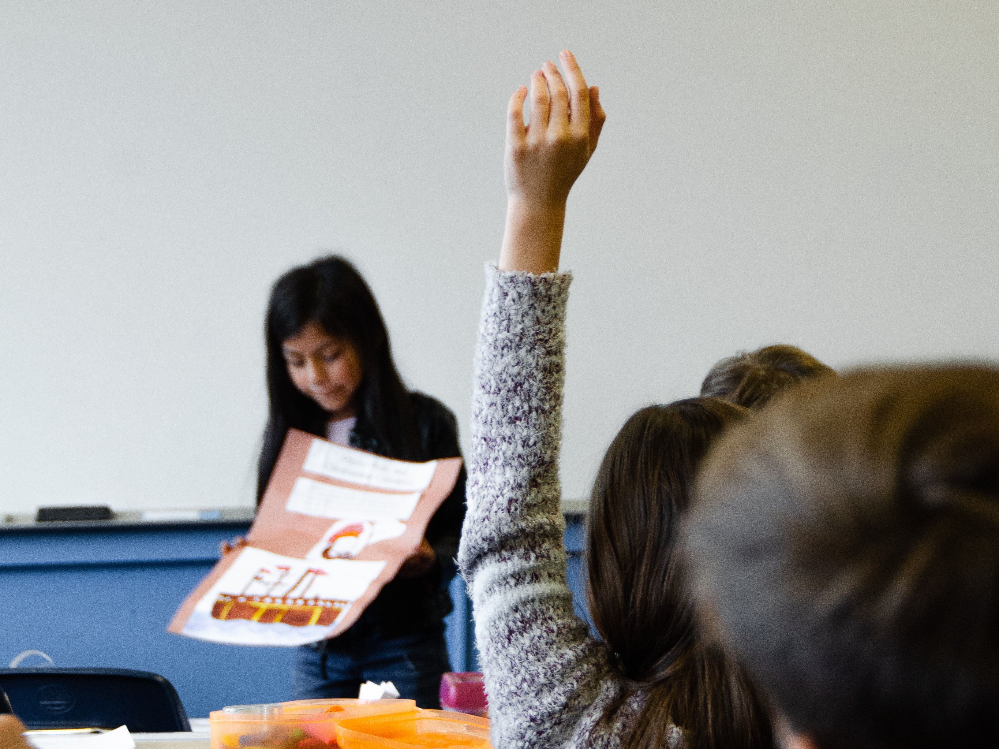 child gives a presentation in front of a classroom, while a fellow student raises their hand