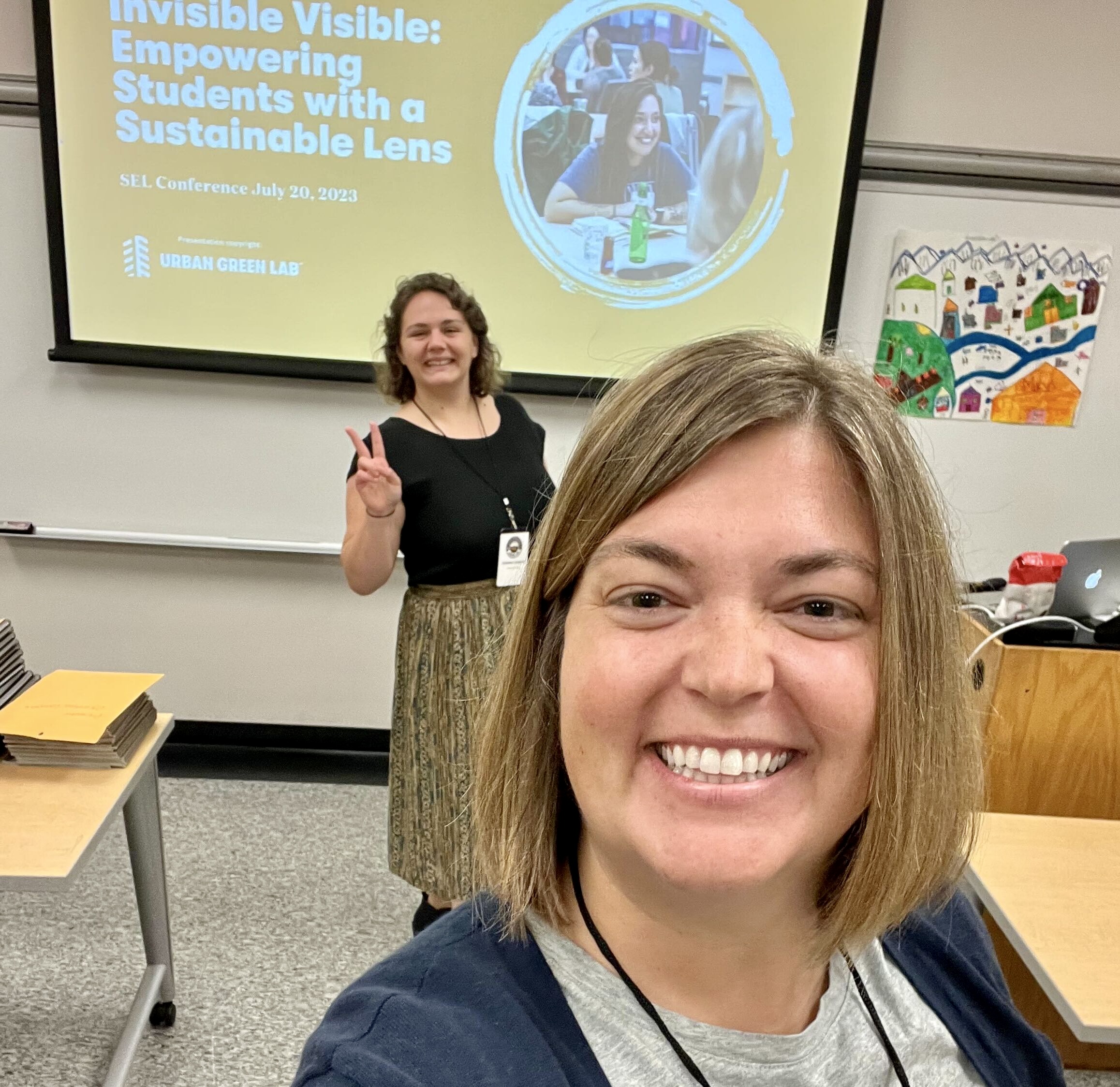 Two women take a selfie in front of a presentation in a classroom