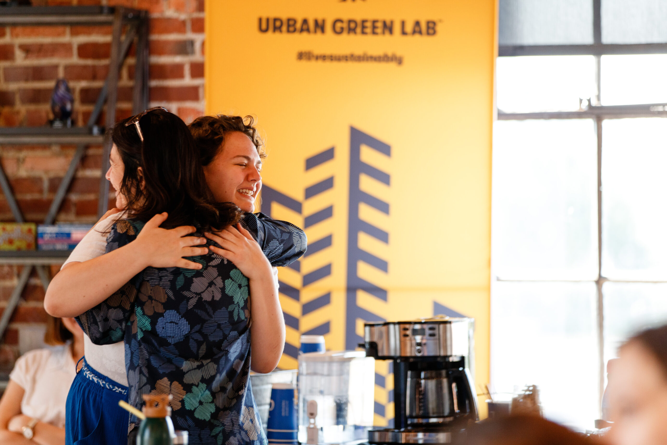 Two women hug in front of an Urban Green Lab sign