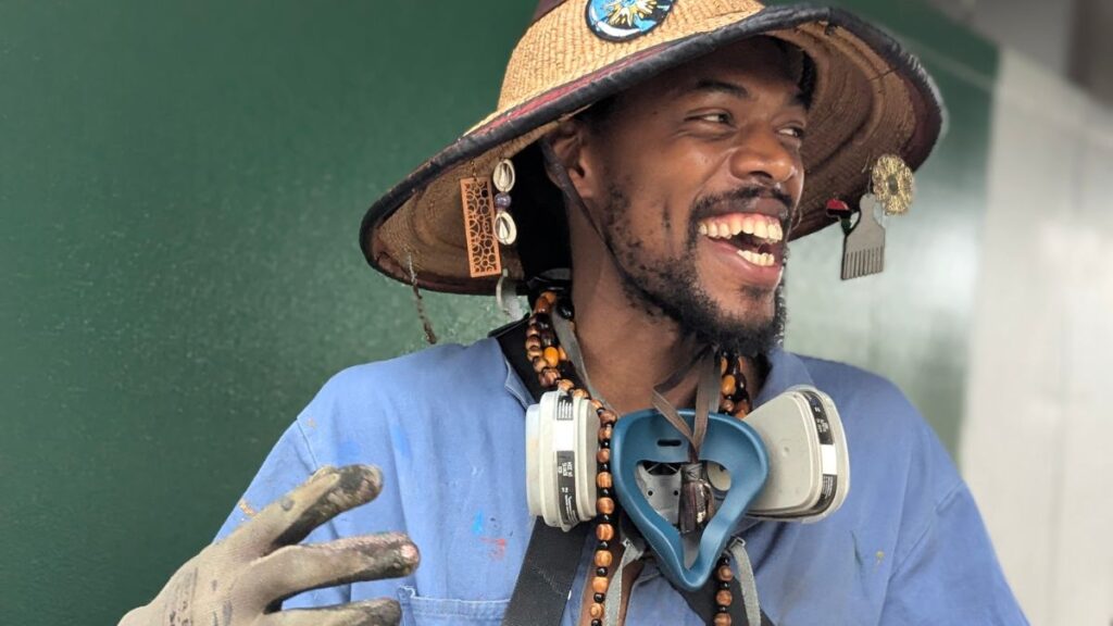 man in a straw hat with a ventilator mask around his neck standing in front of a primed mural wall