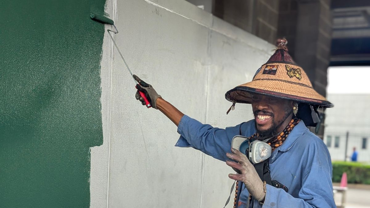 2 artist in straw hat with ventilator mask around his neck runs a paint roller with green paint over a wall