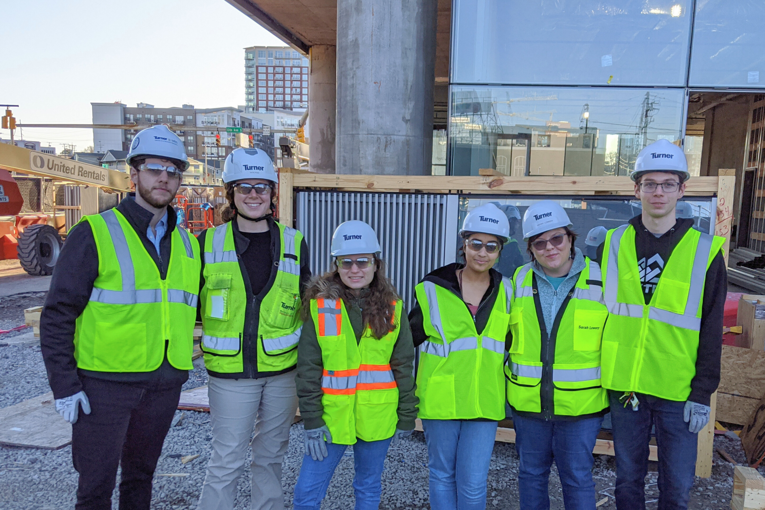 team in safety vests and hardhats smiles in front of glass building on. construction site