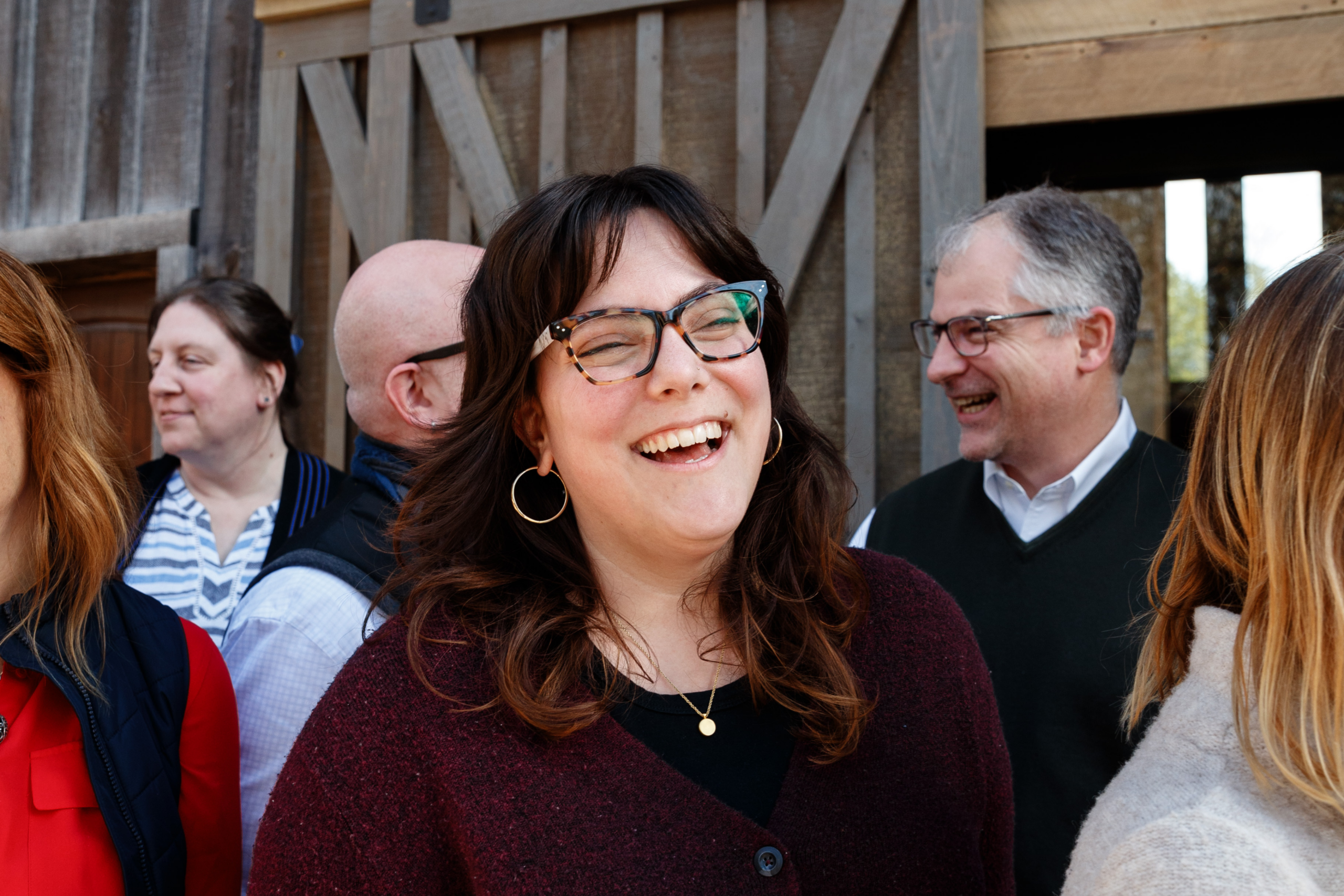 woman with brown hair and glasses smiles in front of a barnwood background with others in the background of the photo
