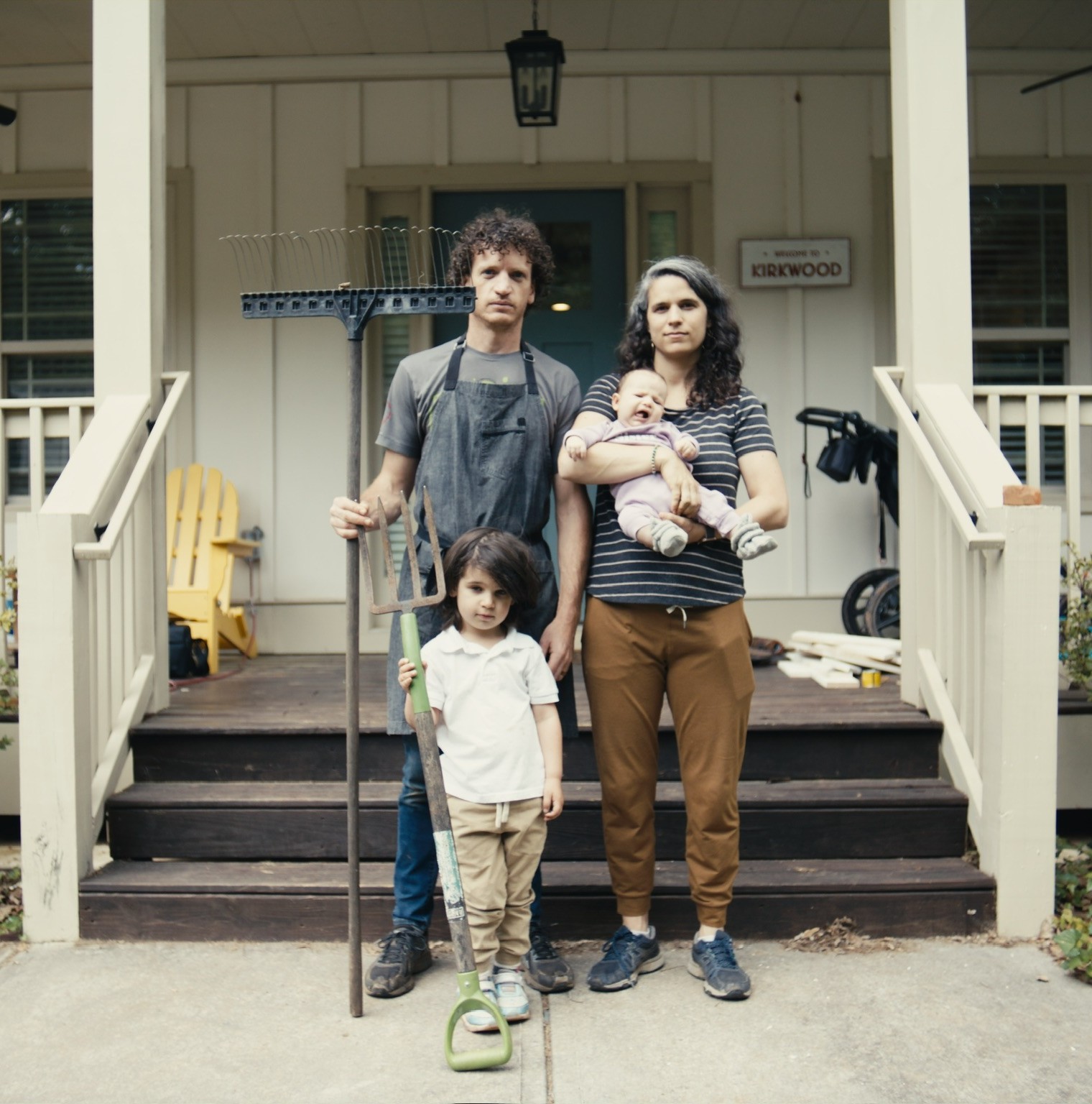 man, woman, and two children positioned in front of a porch with the man holding a rake, in the style of American Gothic