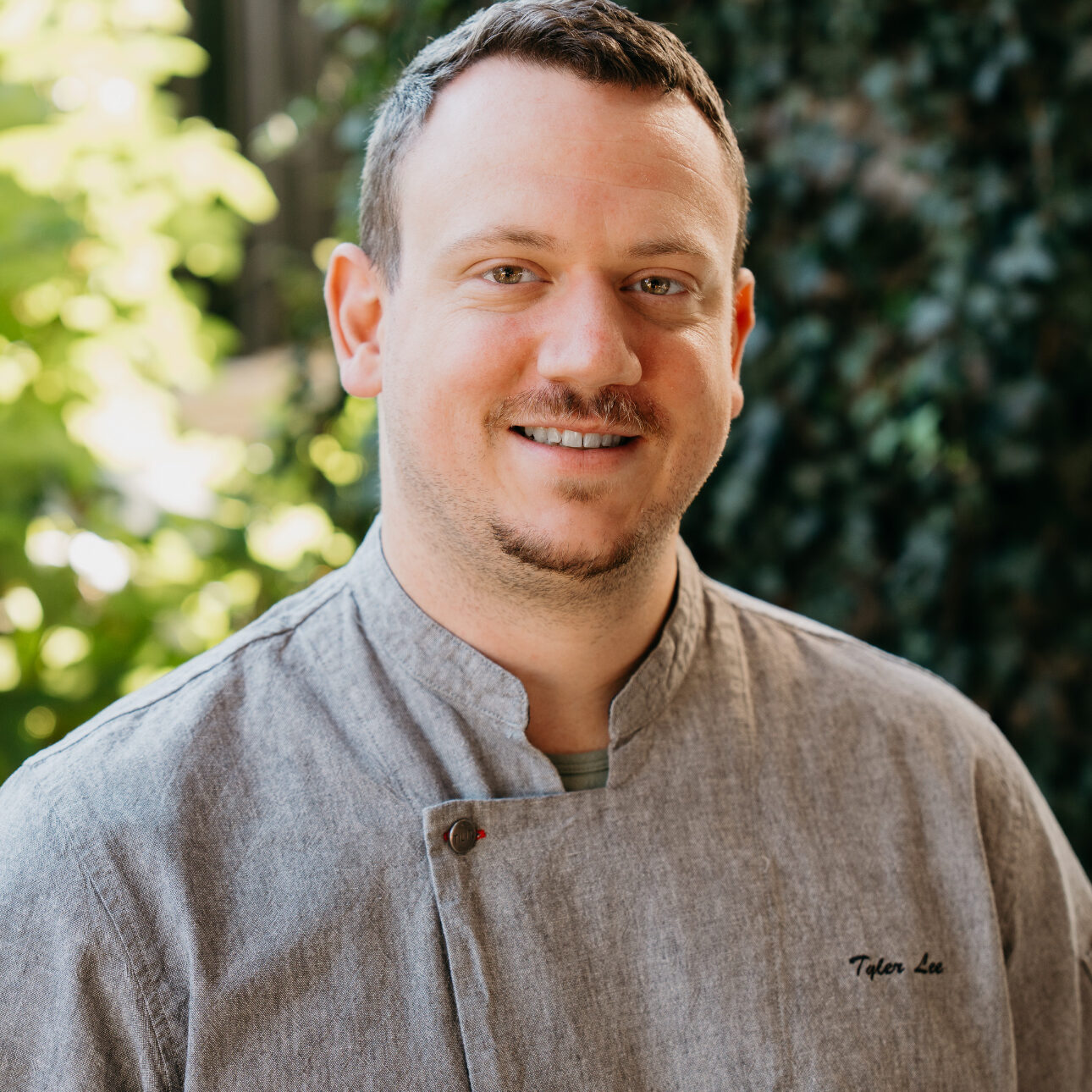 man in a chef's shirt smiles in front of a natural background