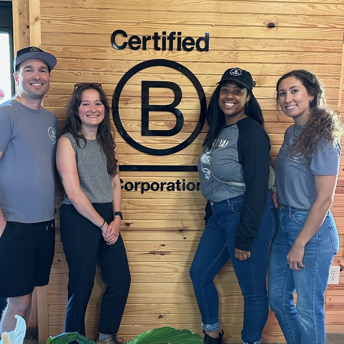 Group of four people pose in front of a B Corp logo on a wood wall