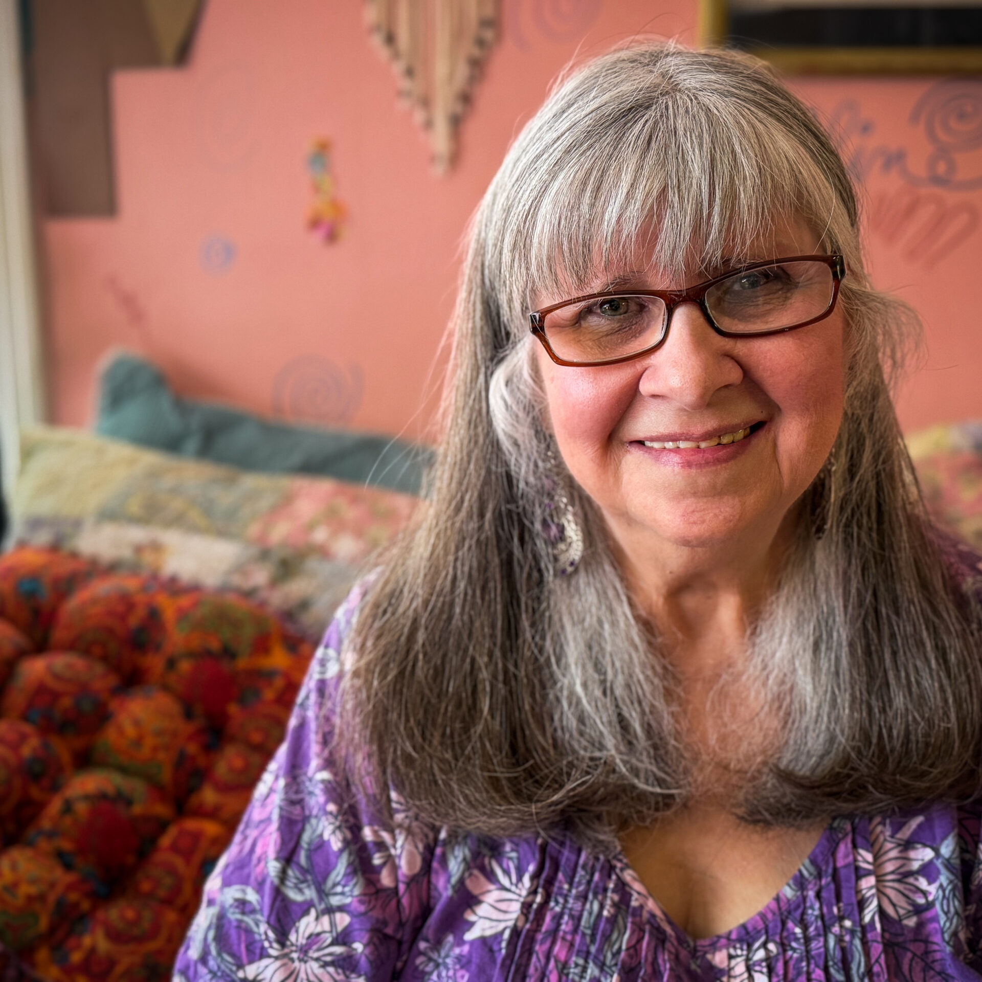 woman with long gray hair and glasses smiles in front of a pink interior wall