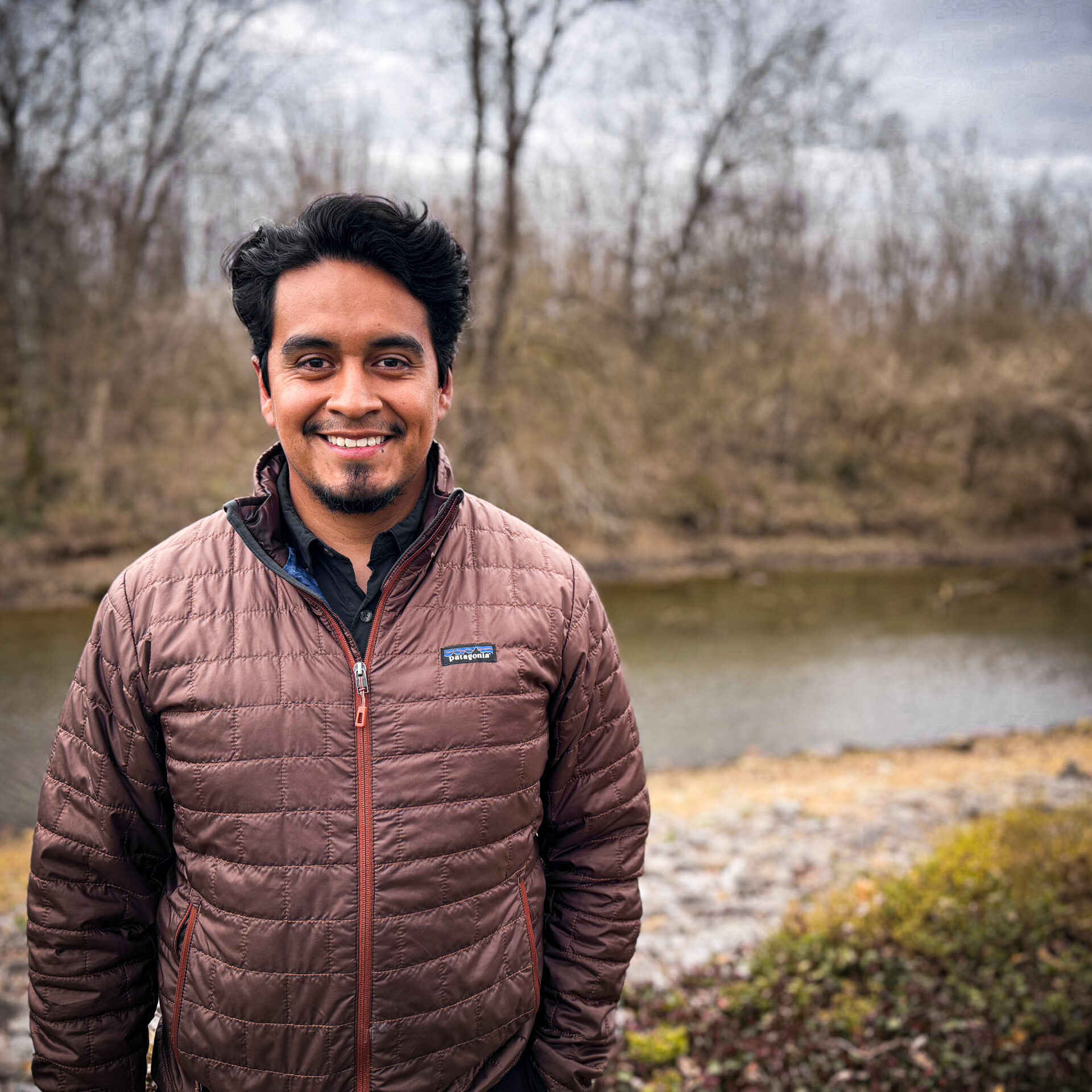 Young dark-haired man in a purple patagonia coat standing in front of a river