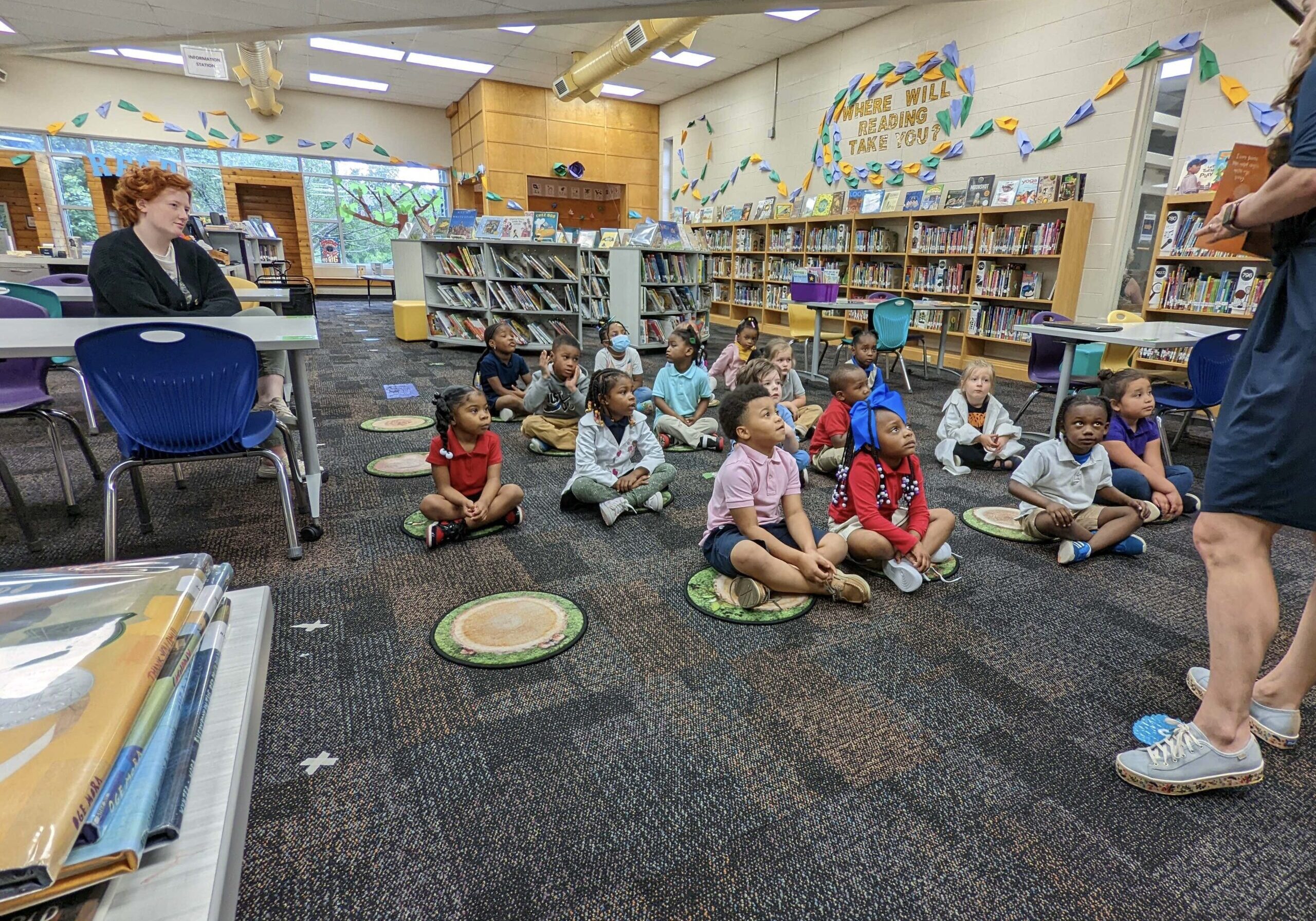 Children sit on the floor in the library while a teacher reads to them