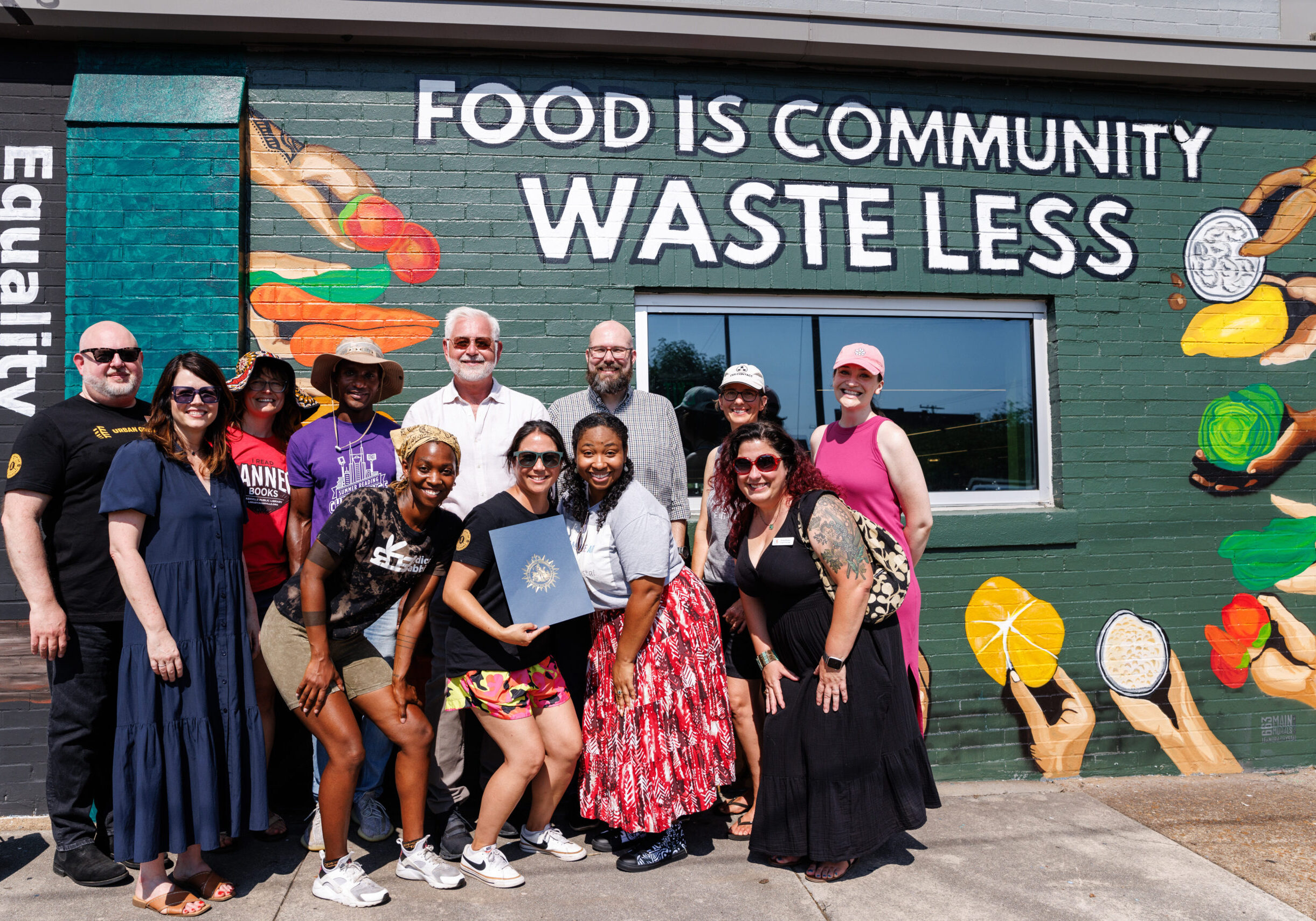 Group of 10 or more people pose for a photo in front of a mural that says "Food is Community. Waste Less."