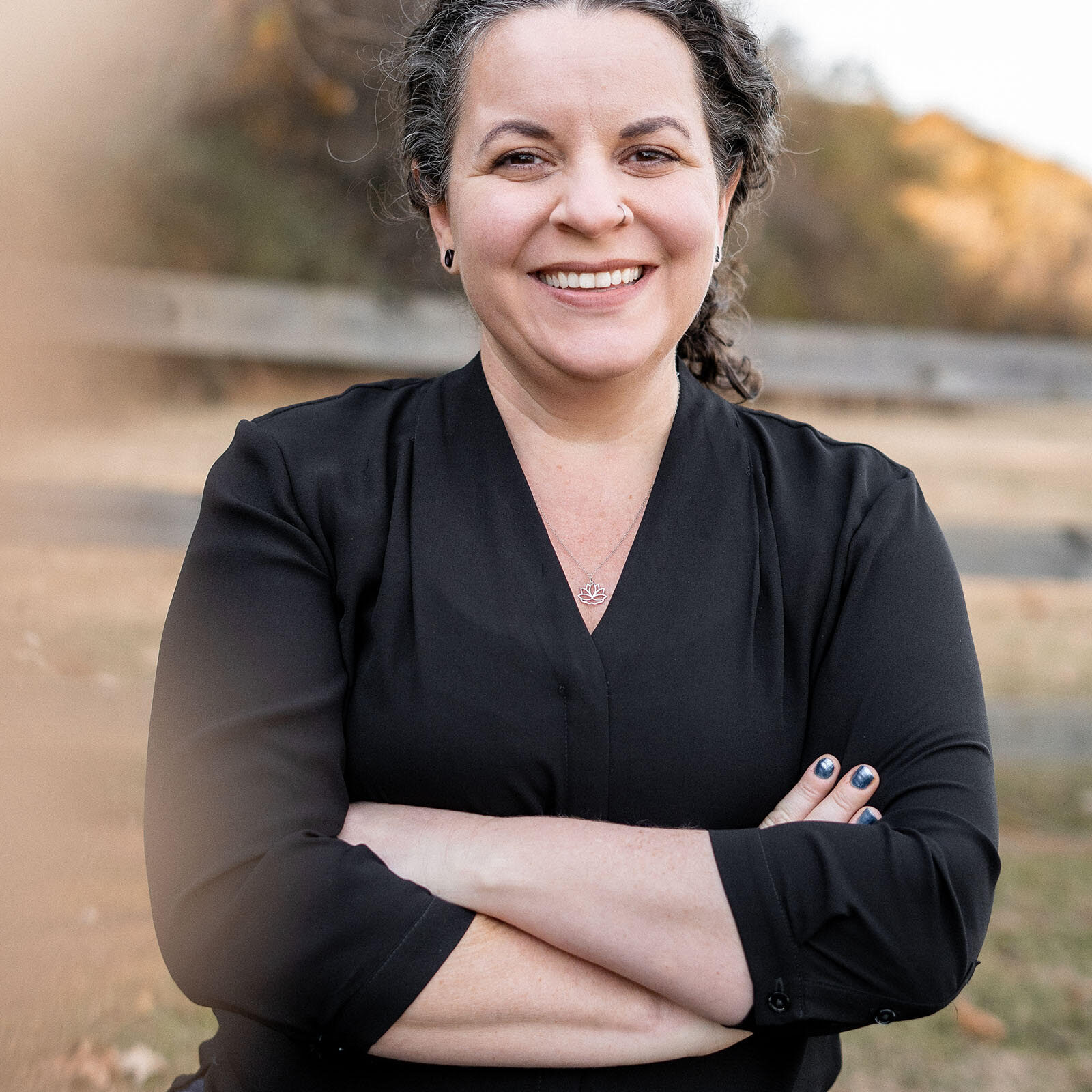 woman in a black blouse smiles with confidently crossed arms in a park with sunset light